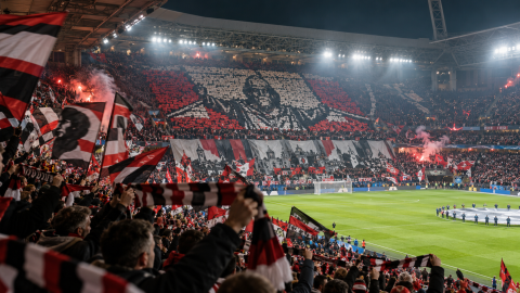 Football supporters displaying a coordinated tifo with flags and scarves across a packed stadium stand