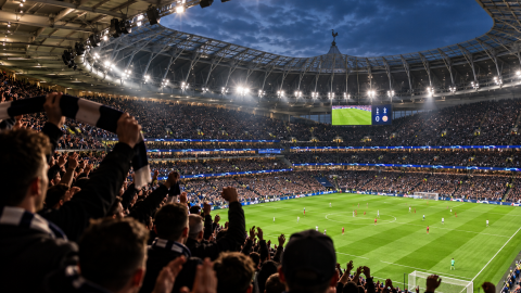 Packed modern football stadium with steep stands, roof structure, floodlights, and supporters surrounding the pitch