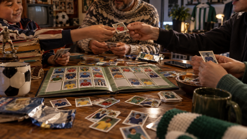 Football fans of different generations trade player stickers around an open sticker album filled with collected and missing football stickers.