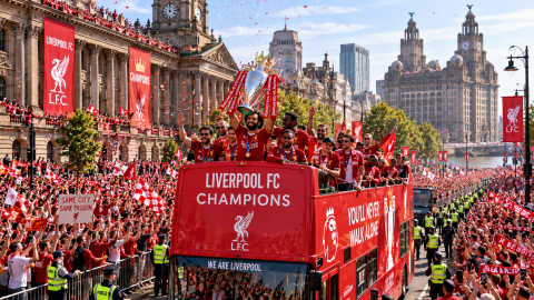 Liverpool FC players celebrating a championship victory on an open-top bus parade, lifting the Premier League trophy as thousands of supporters cheer through the city streets.